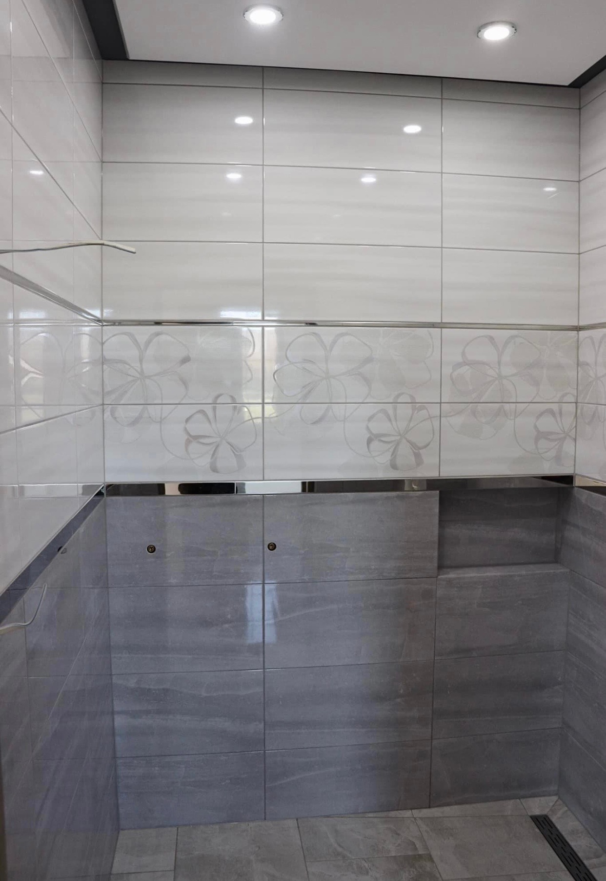 Wykończona ściana kabiny prysznicowej z białymi płytkami z motywem kwiatowym i szarymi płytkami imitującymi kamień, z widocznym odpływem liniowym w podłodze.