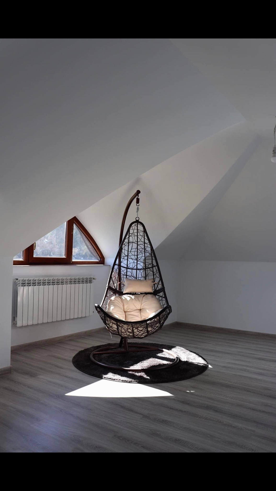 Wiklinowy fotel wiszący z kremowymi poduszkami na okrągłym dywaniku w jasnym pokoju na poddaszu z oknem w kształcie trójkąta.