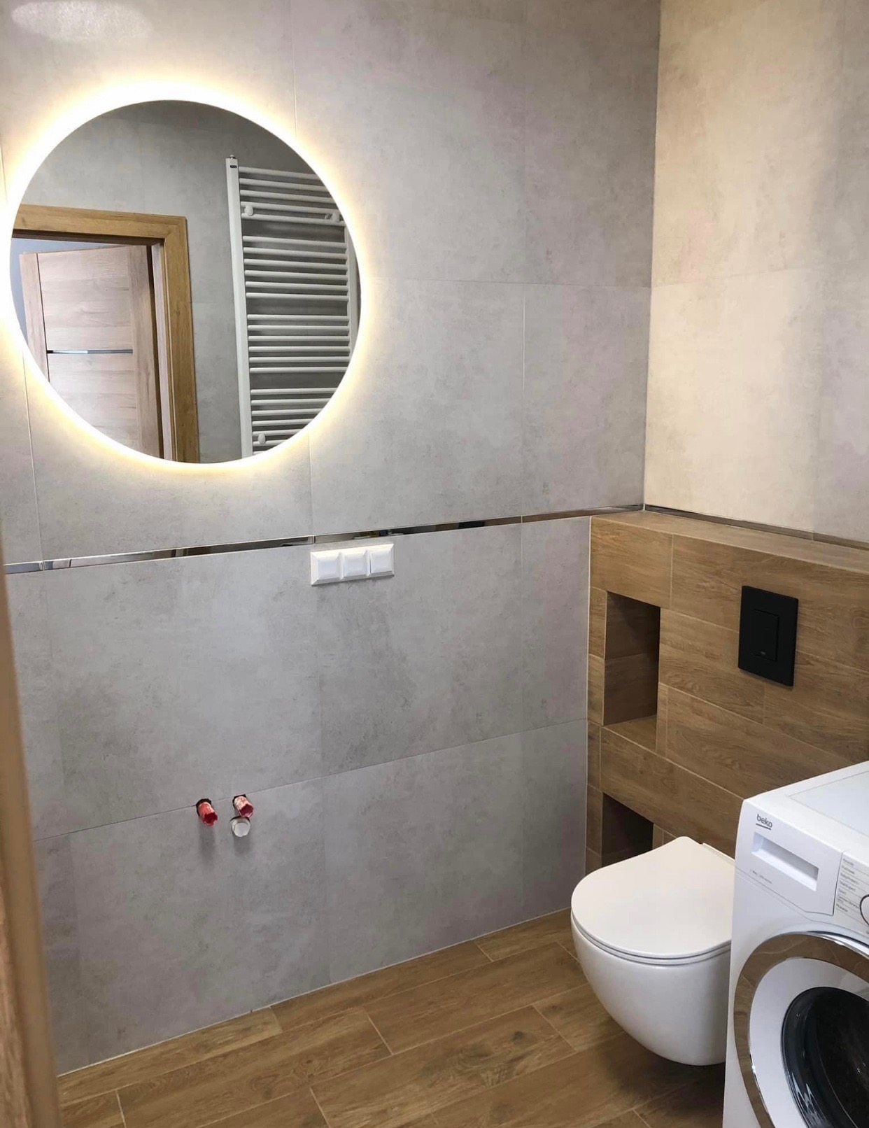 Nowoczesna łazienka z szarymi płytkami, okrągłym lustrem z podświetleniem LED, podwieszaną toaletą i pralką Beko.