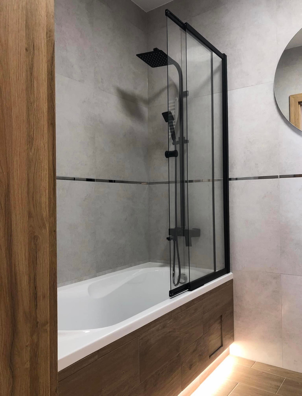 Nowoczesna łazienka z wanną, czarną armaturą i szklanym parawanem nawannowym z czarnymi ramami, oświetlenie LED pod wanną, płytki w odcieniach szarości i beżu.