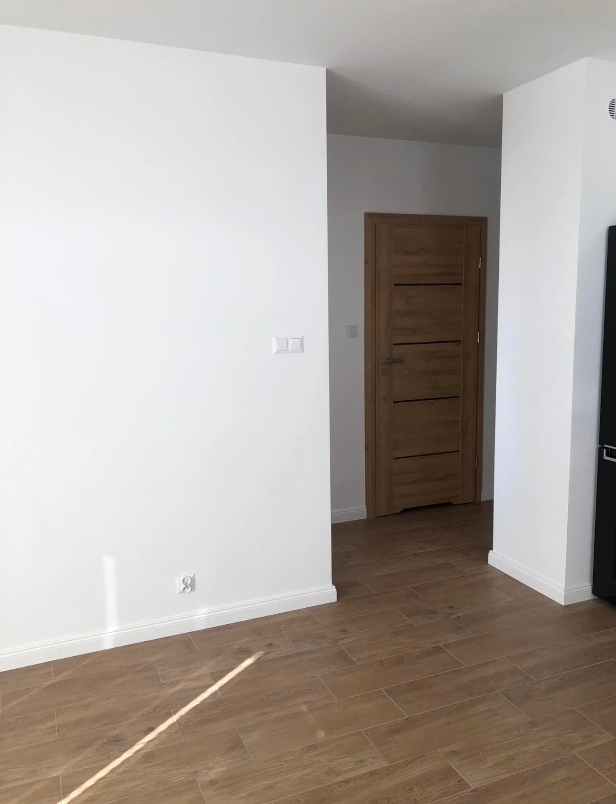 Pusty pokój po remoncie z drewnopodobną podłogą, białe ściany z zamontowanymi kontaktami i włącznikami oraz drewniane drzwi z poziomymi panelami.