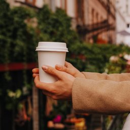 Ph.Radkevych - Dłonie w beżowym płaszczu trzymają biały kubek z kawą na wynos, na tle kamienicy z zielonymi pnączami.