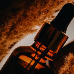 Ph.Radkevych - Amber szklana buteleczka z pipetą na tle puszystych, brązowych traw, z delikatnym światłem podkreślającym fakturę i kształt.