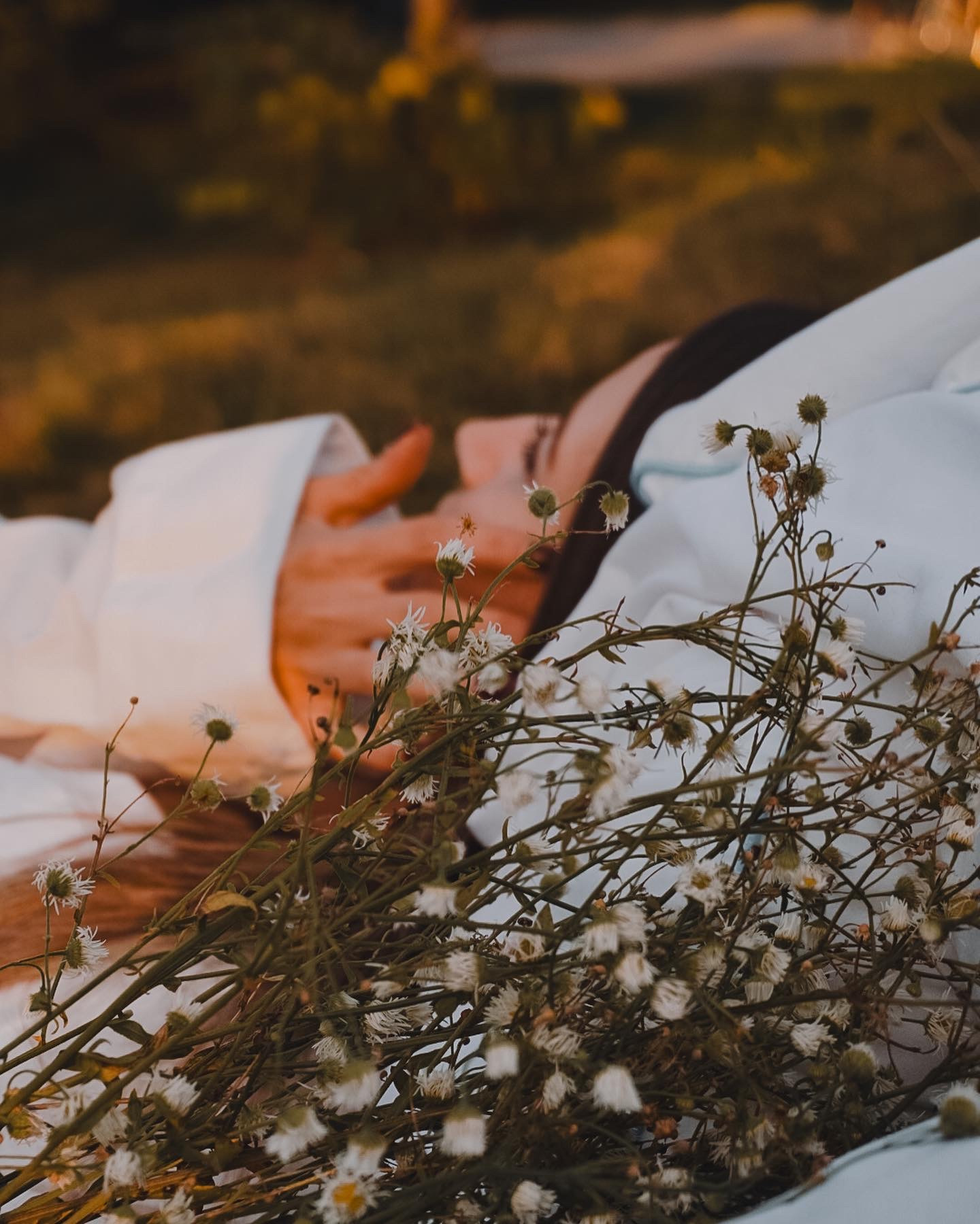 Kobieta w białej koszuli leży na trawie z bukietem polnych kwiatów, delikatne światło zachodzącego słońca.