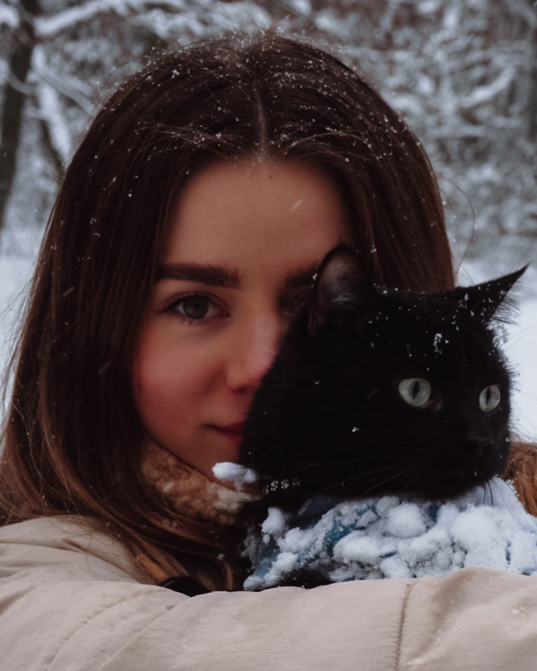 Portret młodej kobiety z czarnym kotem pokrytym śniegiem na tle zimowego krajobrazu.