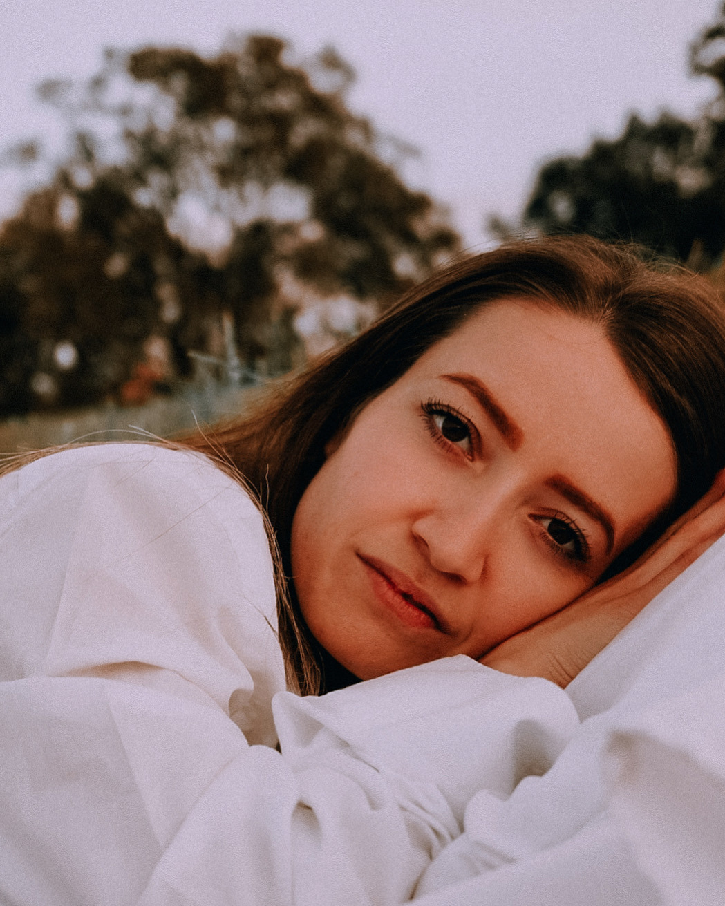 Portret kobiety w białej koszuli, leżącej na jasnym tle z rozmyciem drzew w tle, z delikatnym uśmiechem i spojrzeniem w obiektyw.