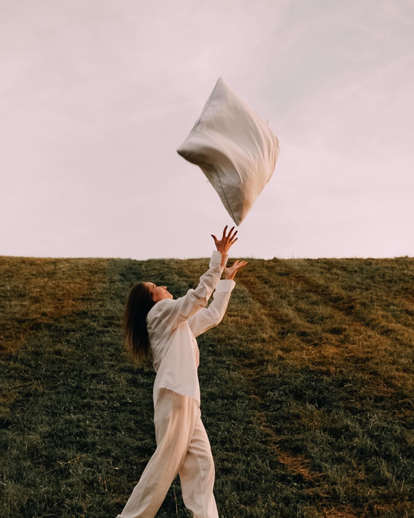 Kobieta w białym stroju rzuca poduszką w górę na tle zielonego wzgórza i jasnego nieba o zachodzie słońca.
