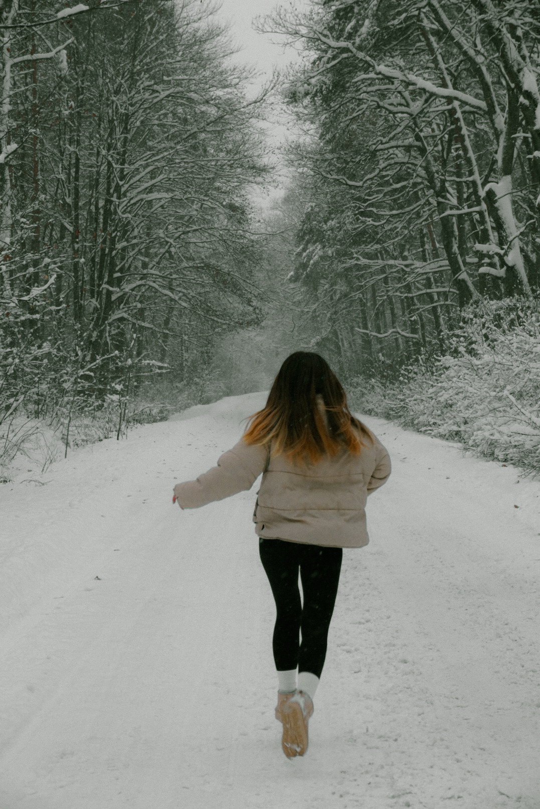Kobieta biegnąca w zimowej scenerii, na śnieżnej drodze w lesie, w beżowej kurtce i czarnych legginsach, z widocznymi blond końcówkami włosów.