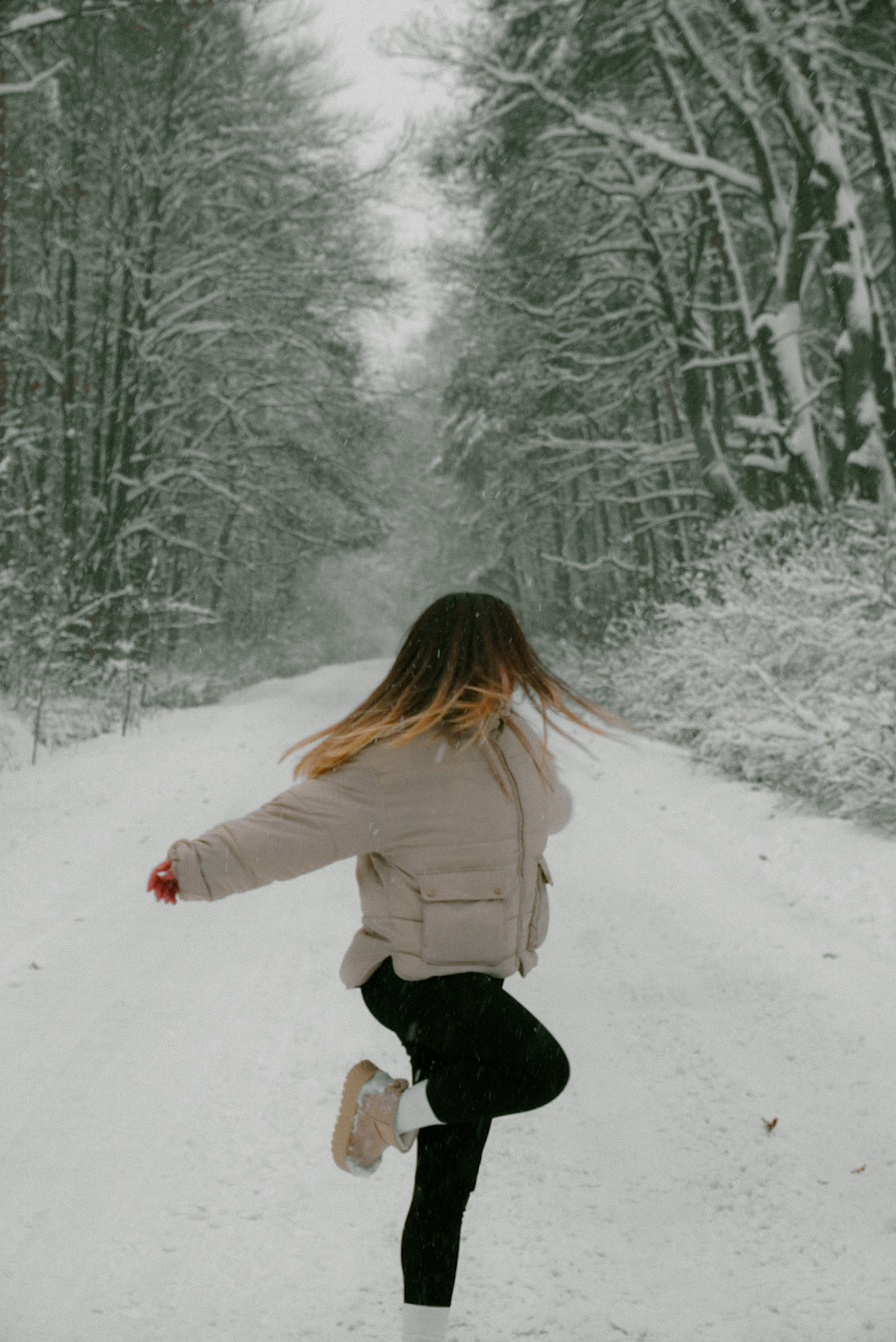 Kobieta w jasnej kurtce i czarnych legginsach tańczy na ośnieżonej drodze otoczonej zimowym lasem, włosy rozwiane, lekko rozmazane od ruchu.