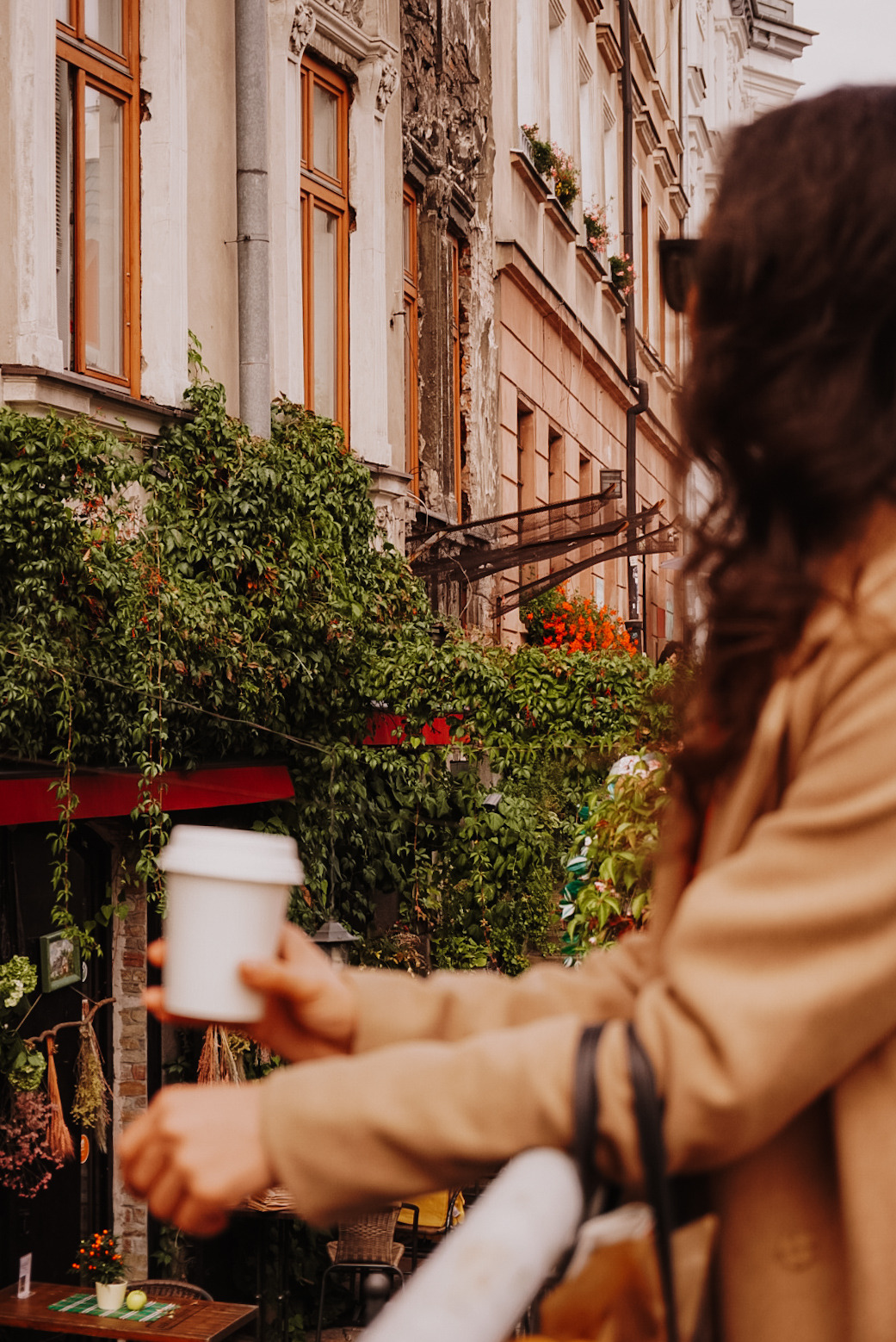 Kobieta z kubkiem kawy na tle kamienicy z bluszczem i kwiatami w oknach.