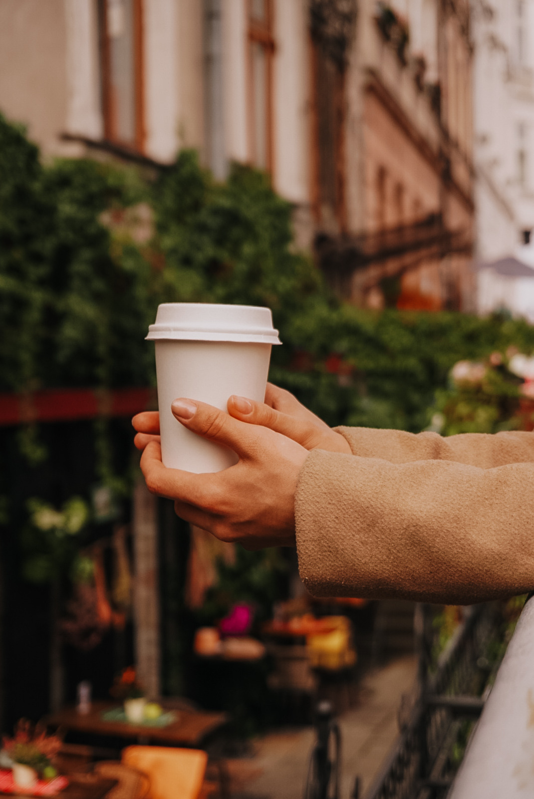 Dłonie w beżowym płaszczu trzymają biały kubek z kawą na wynos, na tle kamienicy z zielonymi pnączami.