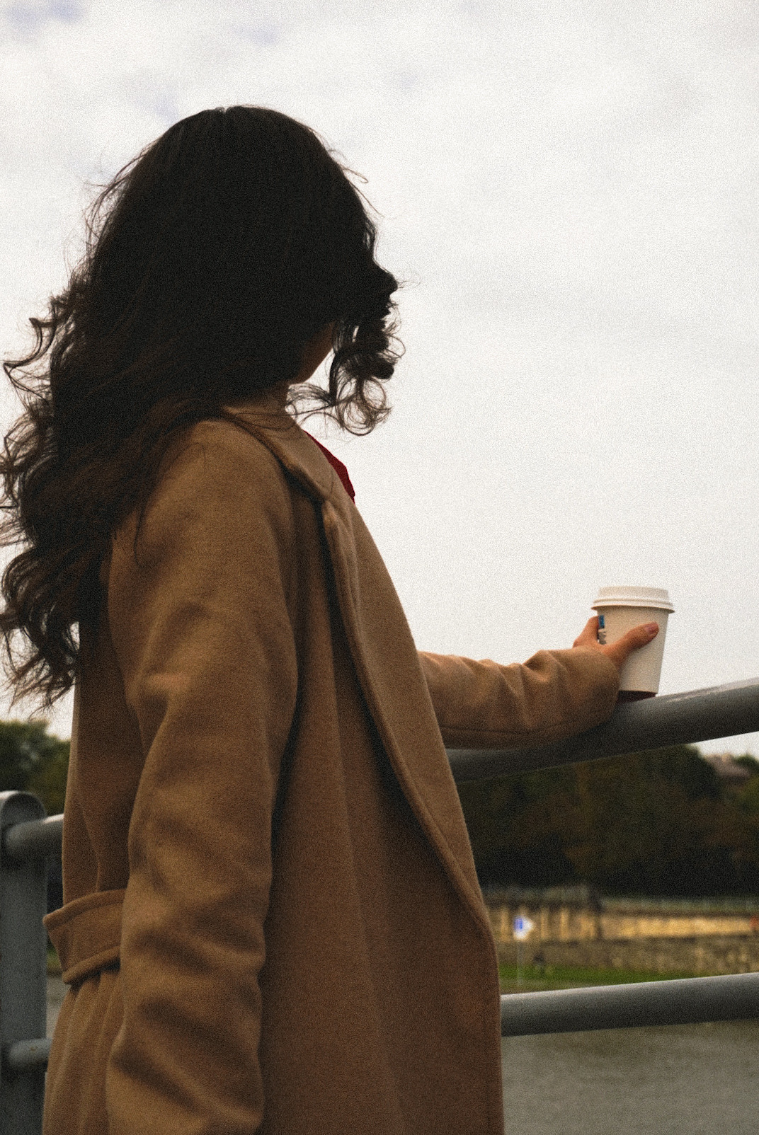 Kobieta w płaszczu z kubkiem kawy, oparta o barierkę z widokiem na rzekę i drzewa w tle.