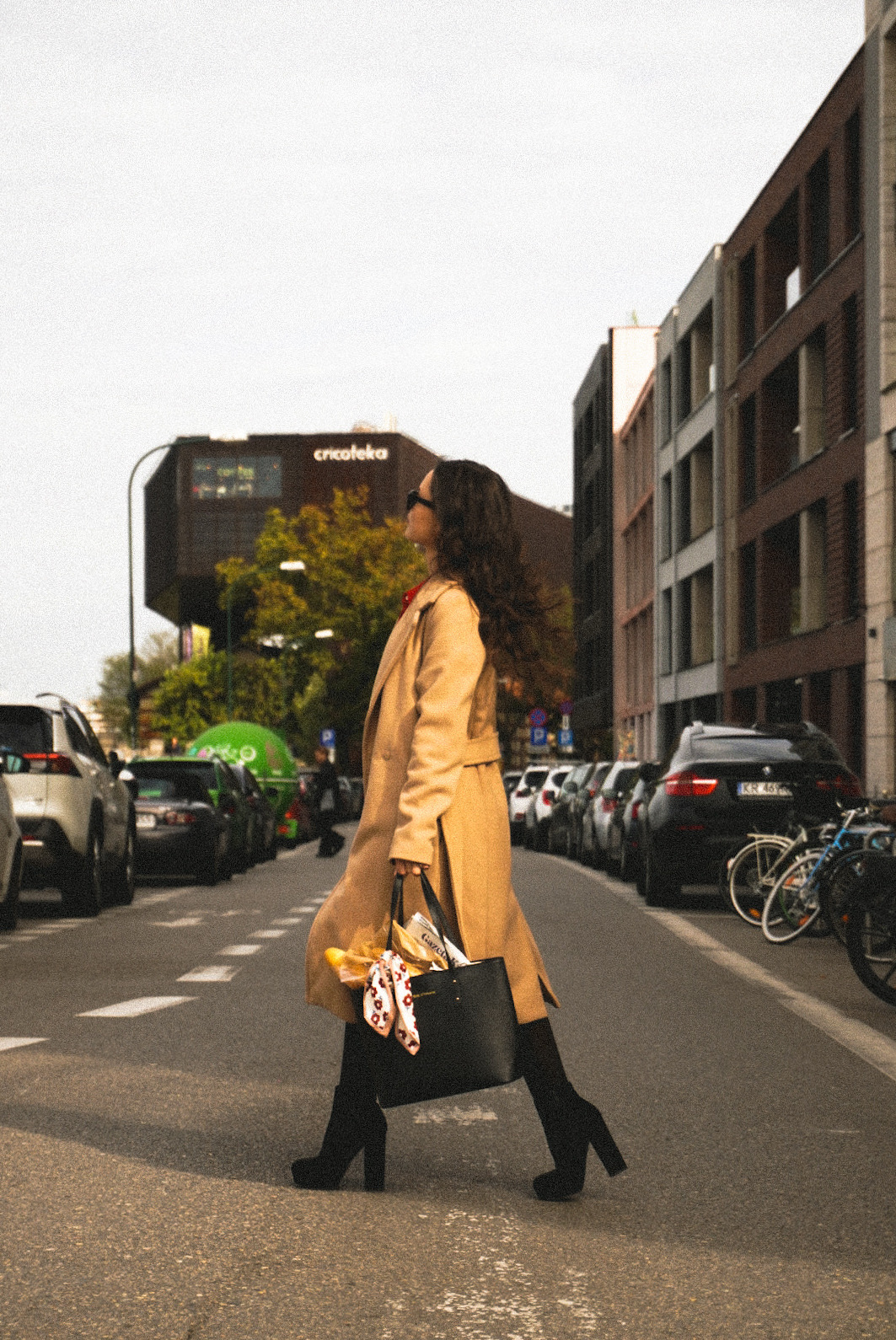 Kobieta w płaszczu przechodzi przez ulicę w mieście, niosąc czarną torbę z bagietką i gazetą.