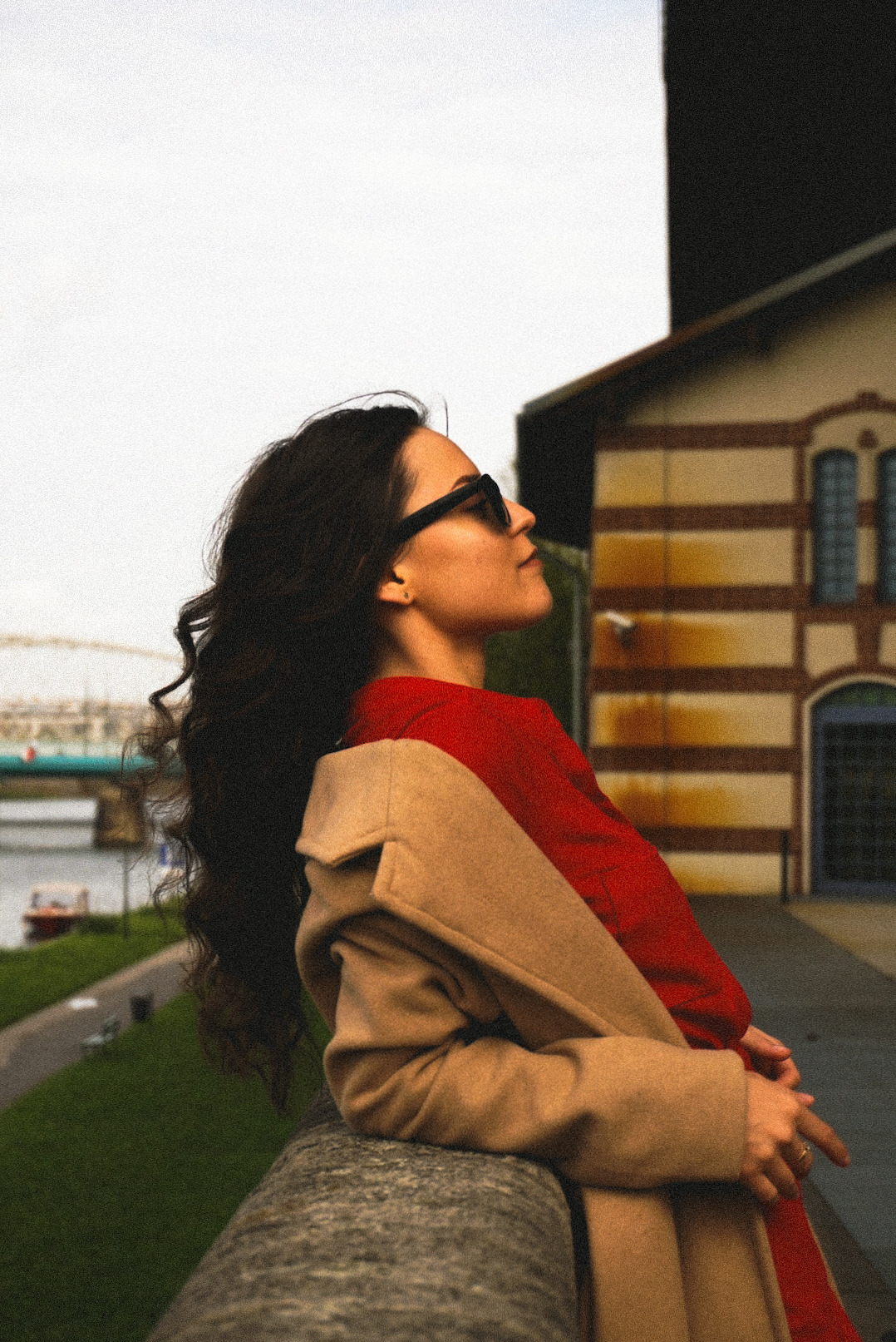 Kobieta w okularach przeciwsłonecznych, czerwonej bluzce i beżowym płaszczu opiera się o kamienną barierkę, spoglądając w górę na tle budynku z cegły i mostu w oddali.