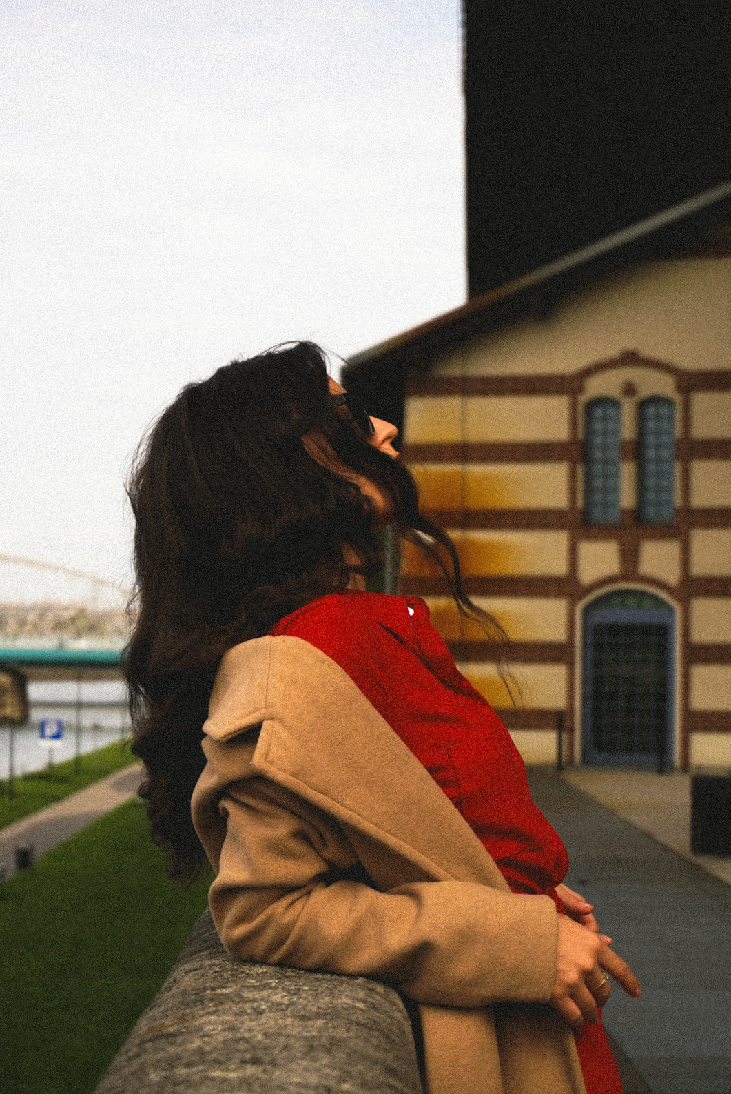 Kobieta w czerwonej koszuli i beżowym płaszczu, odchylona do tyłu, z budynkiem w tle