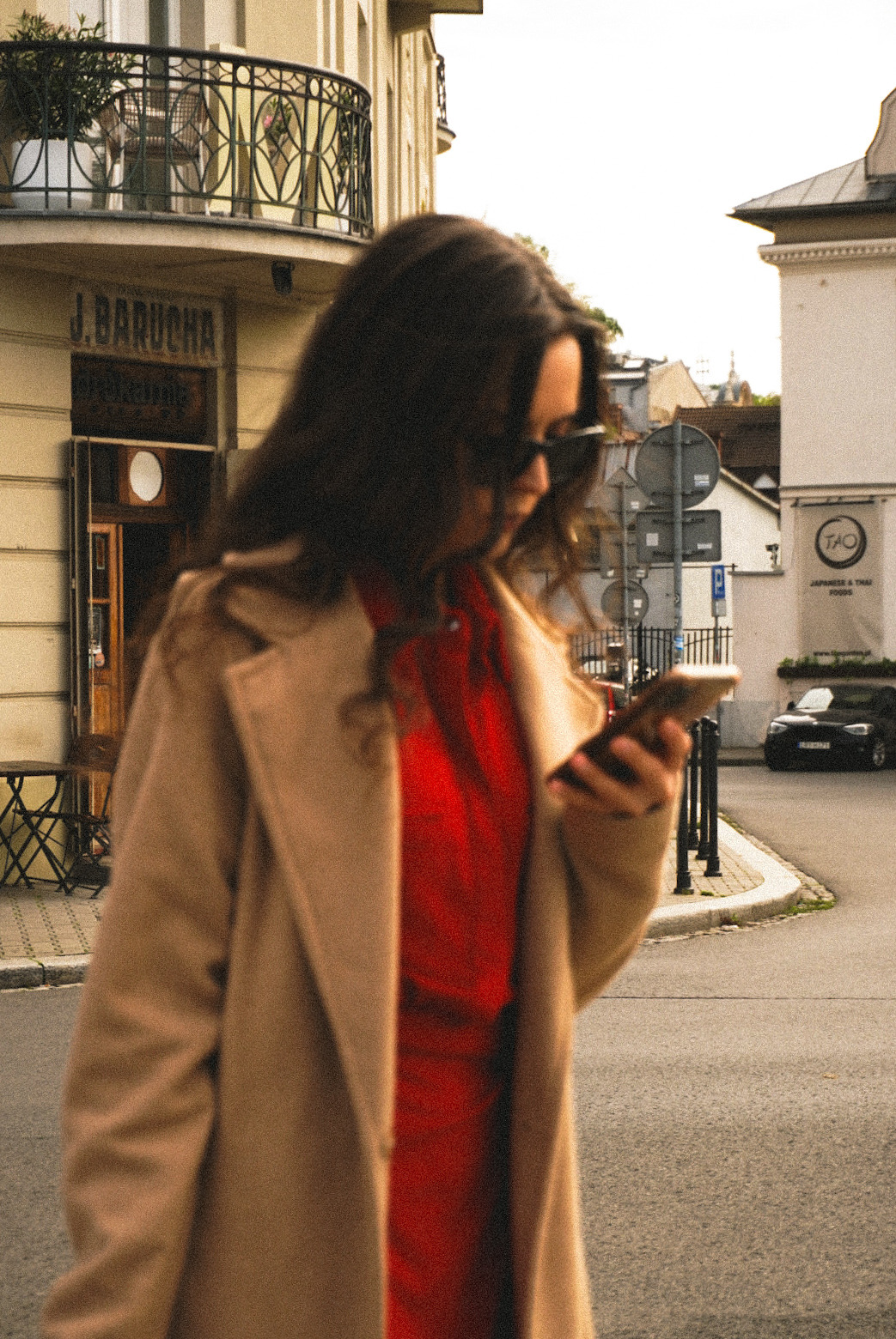 Kobieta w okularach przeciwsłonecznych i beżowym płaszczu sprawdza telefon na tle miejskiej ulicy z budynkami w ciepłych odcieniach i szyldem 'J. BARUCHA'.