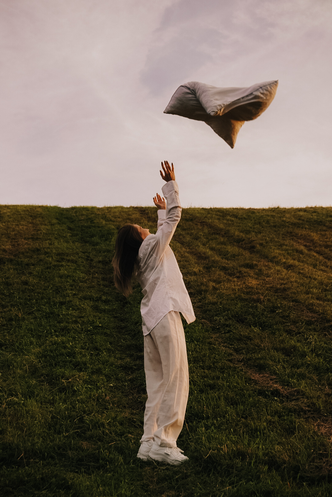 Kobieta w białym, lnianym ubraniu rzuca poduszką w górę na tle zielonego zbocza i zachodzącego słońca.