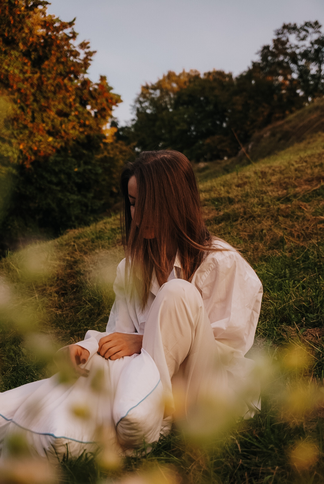 Kobieta w białym stroju siedzi na trawiastym wzgórzu, jesienne drzewa w tle, ujęcie przez rozmyte kwiaty na pierwszym planie.