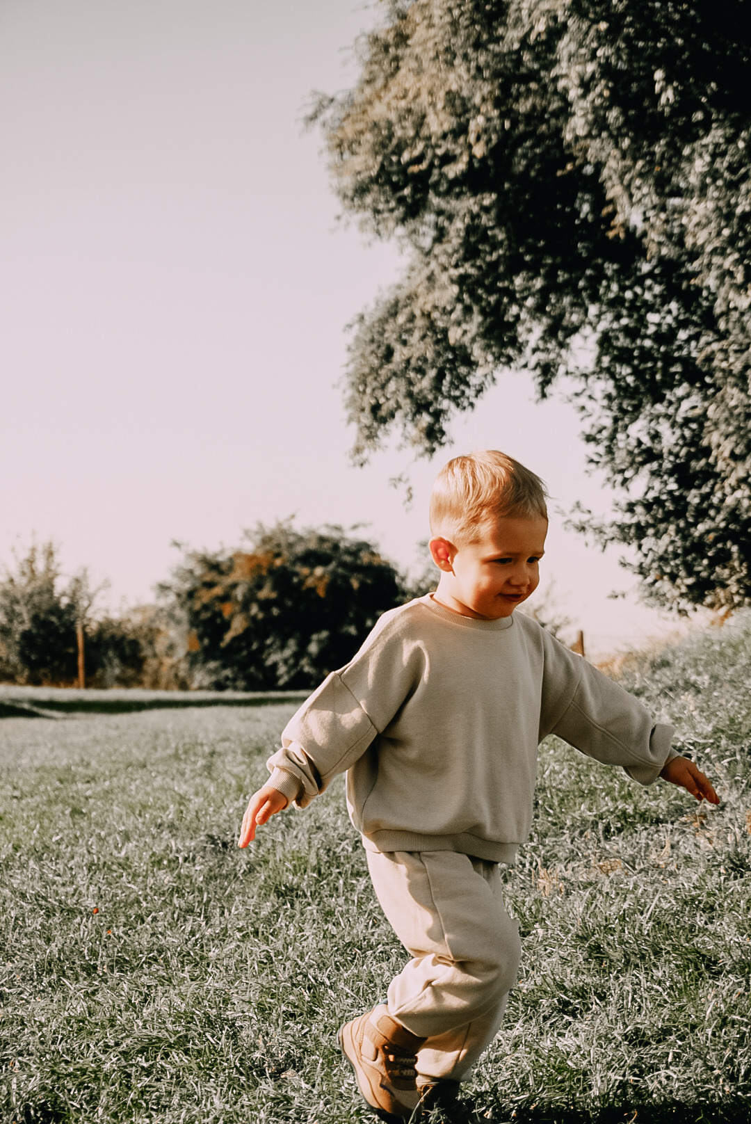 Chłopiec z blond włosami biegnie po trawie w beżowym dresie i brązowych butach, z rozłożonymi ramionami, w tle drzewa i jasne niebo.