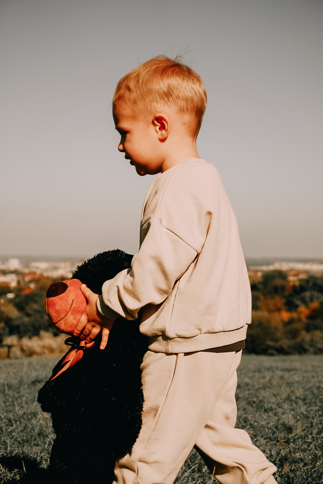 Chłopiec w beżowym dresie trzyma pluszowego misia z czerwoną głową na tle pagórkowatego krajobrazu i jasnego nieba.