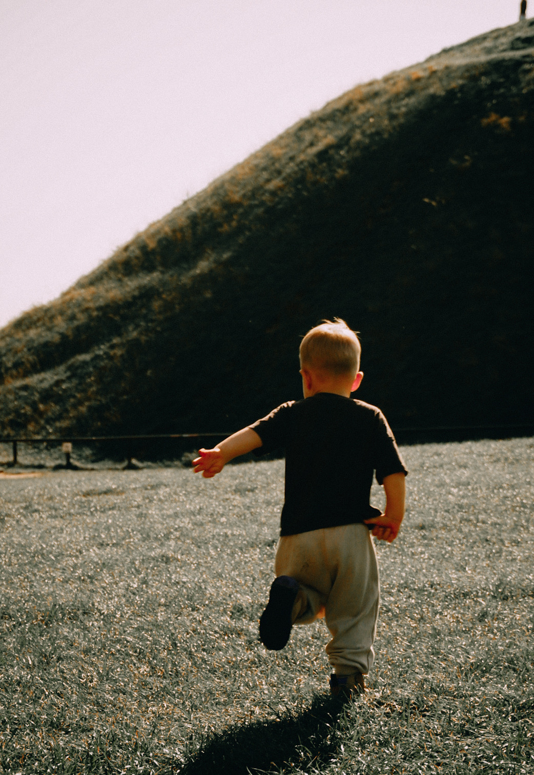 Chłopiec biegnie po trawie w kierunku ciemnego wzgórza, widok z tyłu, jedno ramię wyciągnięte, w tle widoczna sylwetka osoby na szczycie wzniesienia.