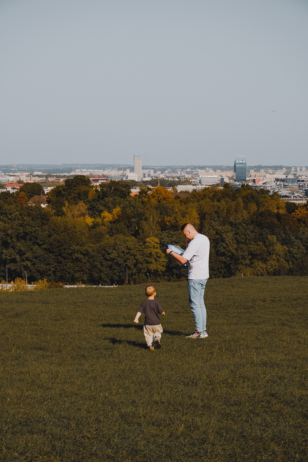 Ojciec przegląda zdjęcia na tablecie, a jego mały syn biegnie po trawie w parku w Krakowie, w tle panorama miasta.