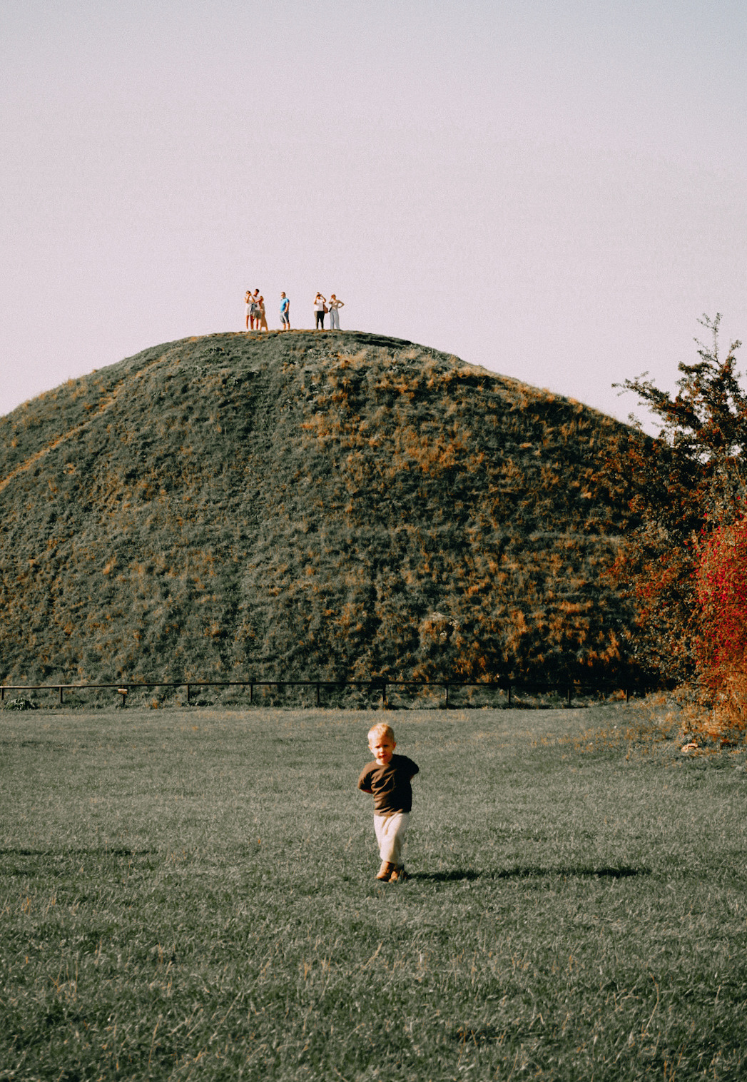 Chłopiec w ciemnej koszulce i jasnych spodniach biegnie po trawie, w tle kopiec z grupą ludzi na szczycie, jesienne drzewo po prawej.