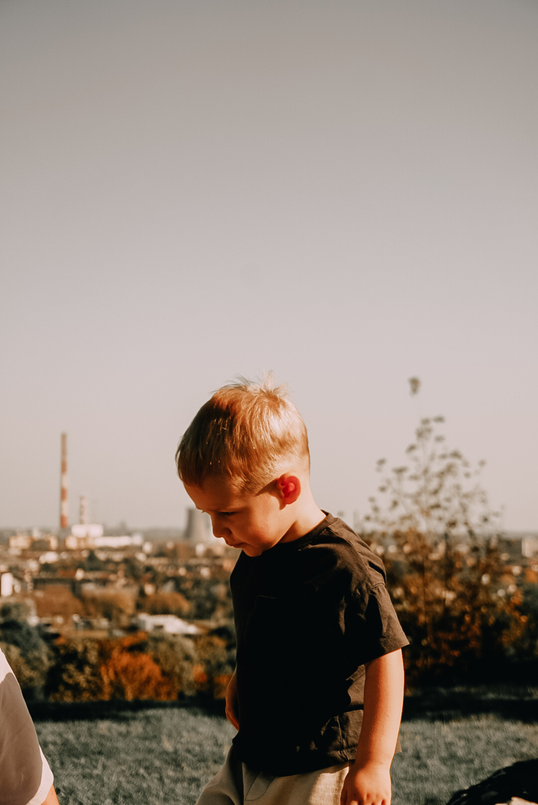 Portret skupionego, jasnowłosego chłopca z profilu, na tle panoramy miasta z kominami fabrycznymi i jesienną roślinnością.