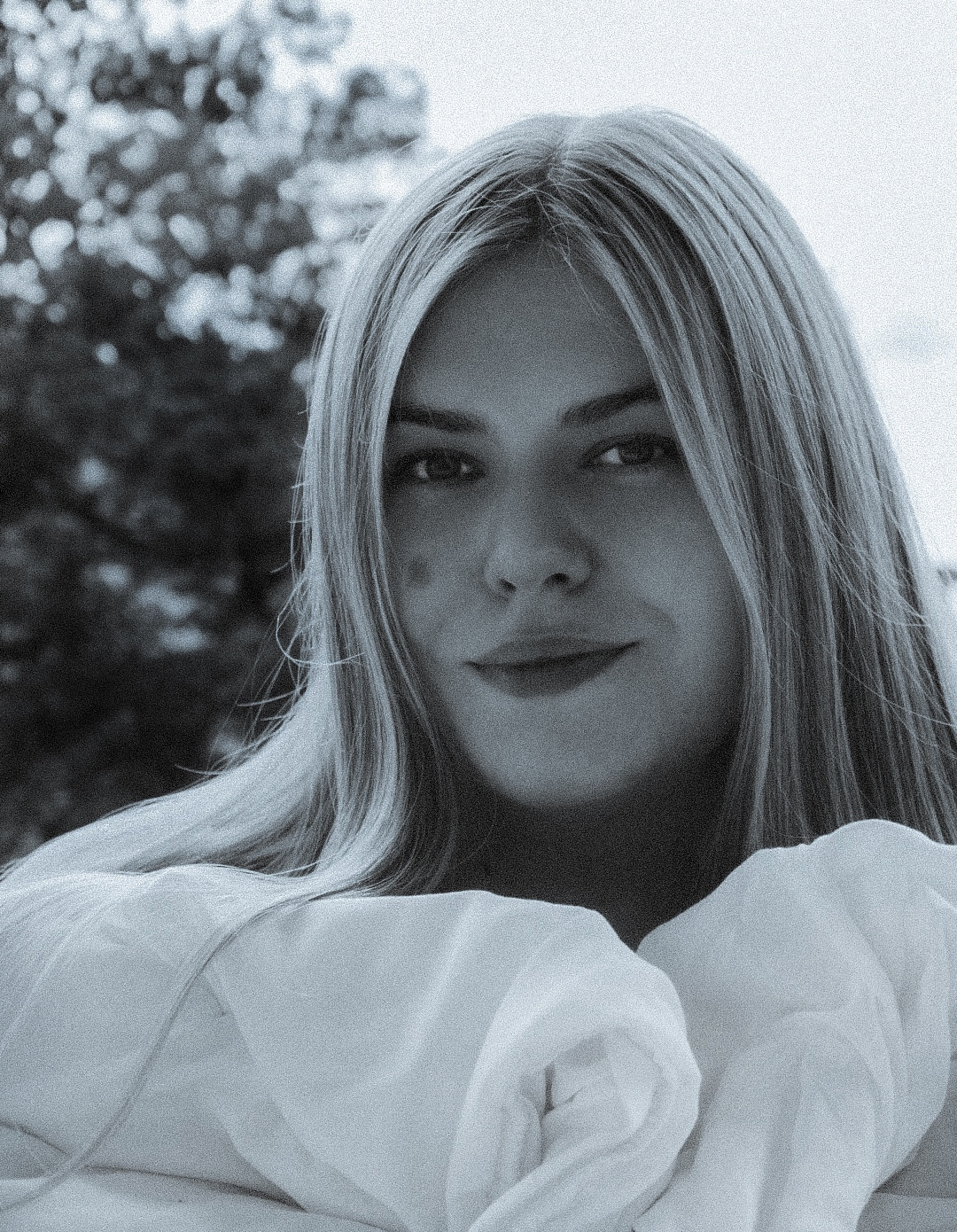 Portret młodej kobiety o jasnych włosach, w białej bluzce, z delikatnym uśmiechem, na rozmytym tle zieleni.