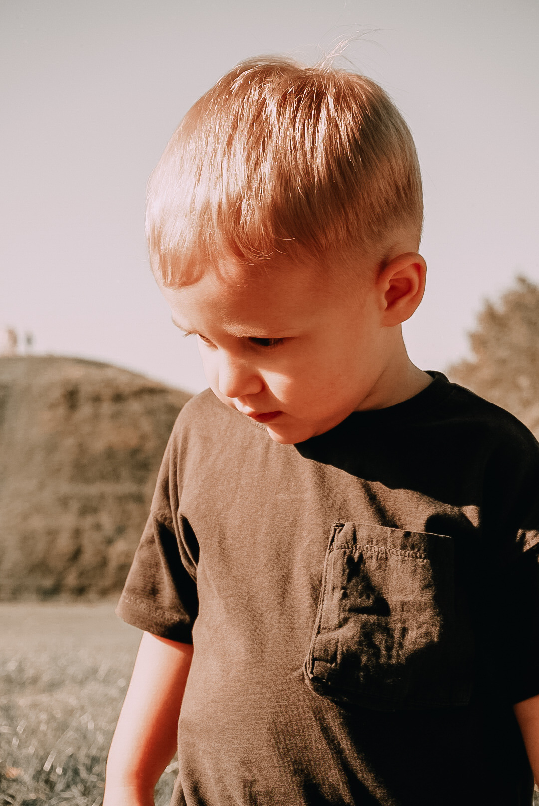 Portret zamyślonego, rudowłosego chłopca w brązowej koszulce z kieszonką, na tle pagórkowatego krajobrazu w ciepłych, stonowanych barwach.