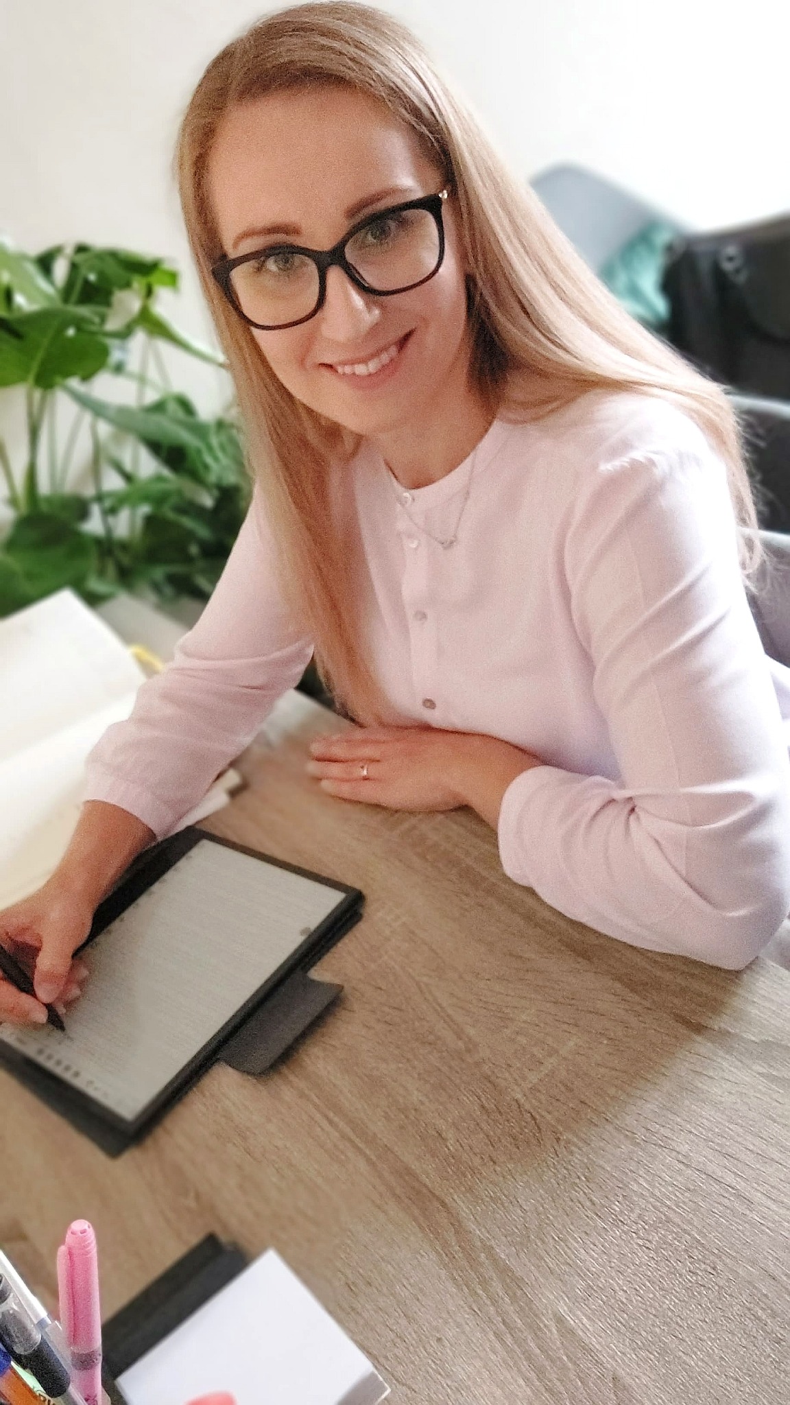 Kobieta w okularach, uśmiechnięta, w jasnoróżowej bluzce, pisze na tablecie graficznym przy biurku, w tle widoczna roślina doniczkowa.