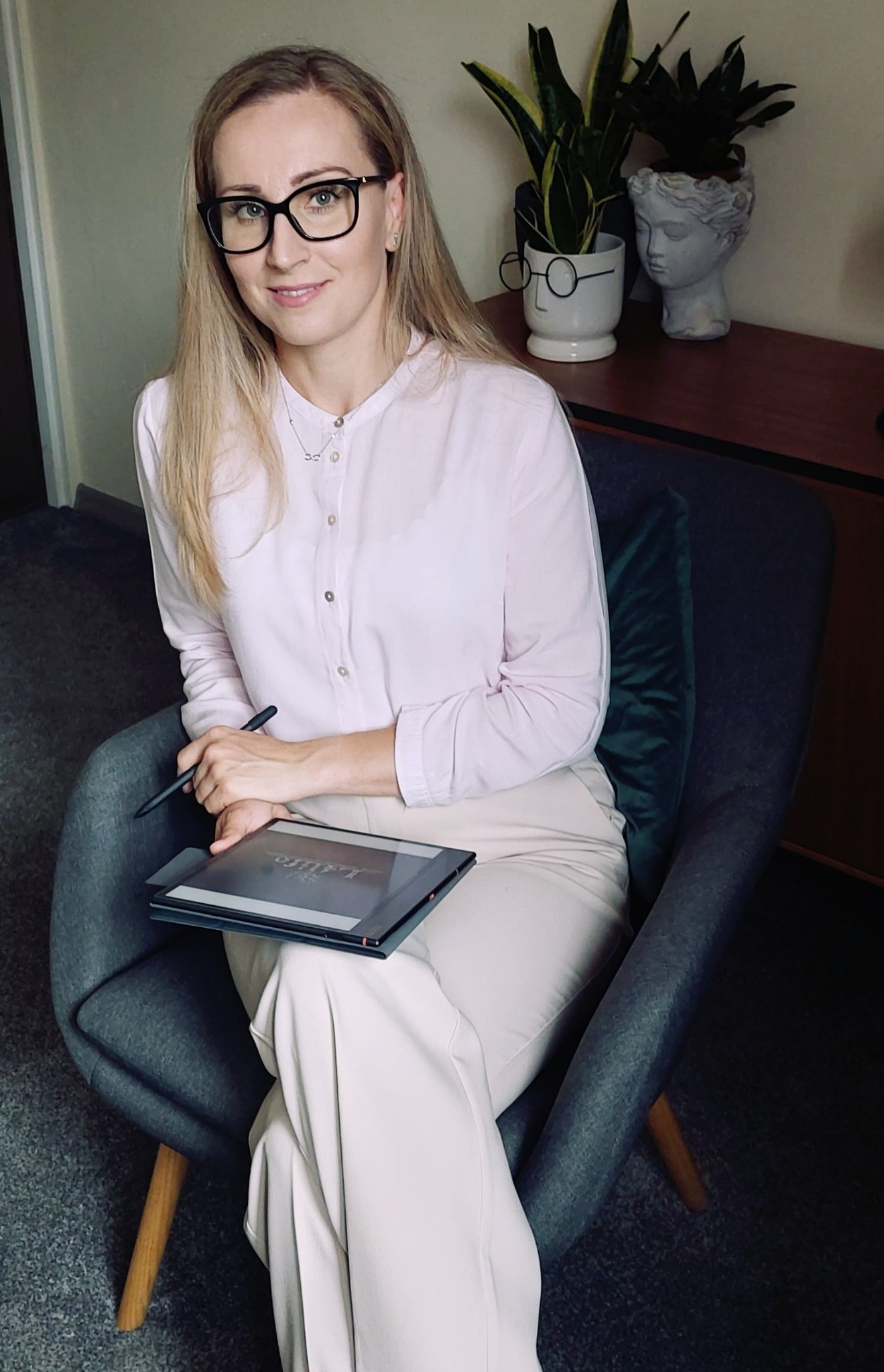 Kobieta w okularach siedzi w fotelu z tabletem graficznym, w tle roślina w doniczce w kształcie głowy i druga roślina w białej doniczce z okularami.