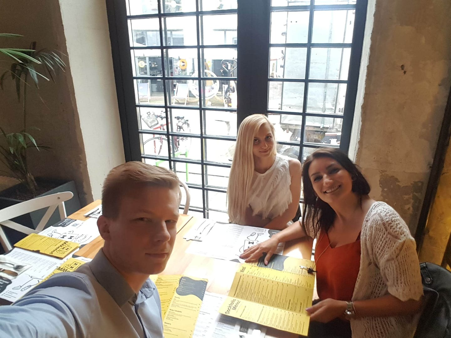 Trzy osoby przeglądają dokumenty na stole w kawiarni, z widokiem na ulicę przez okno w tle.