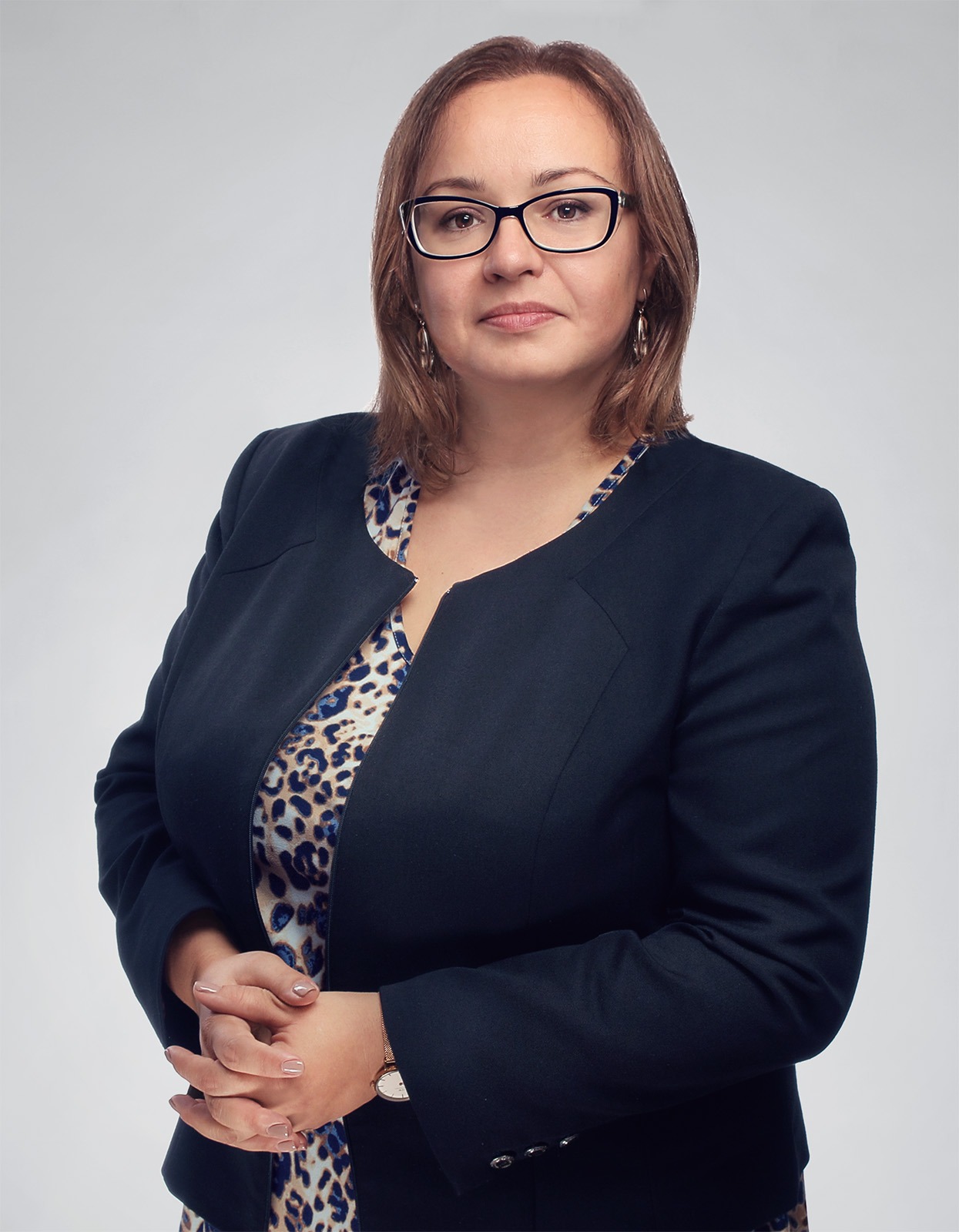 Portret kobiety w okularach, w ciemnej marynarce i bluzce w panterkę, z rękami splecionymi przed sobą, patrzącej wprost w obiektyw na jasnoszarym tle.
