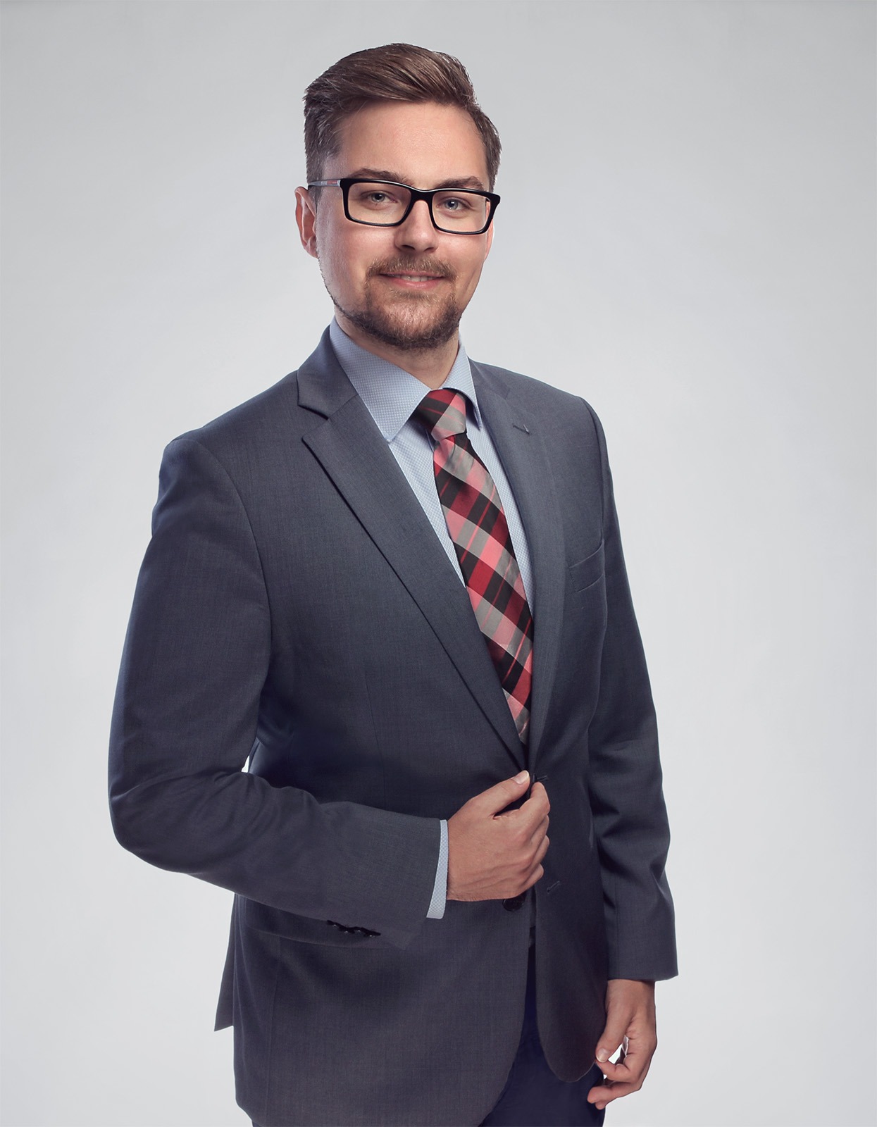 Portret mężczyzny w szarym garniturze, okularach i krawacie w czerwoną kratę, pozującego na jasnoszarym tle.