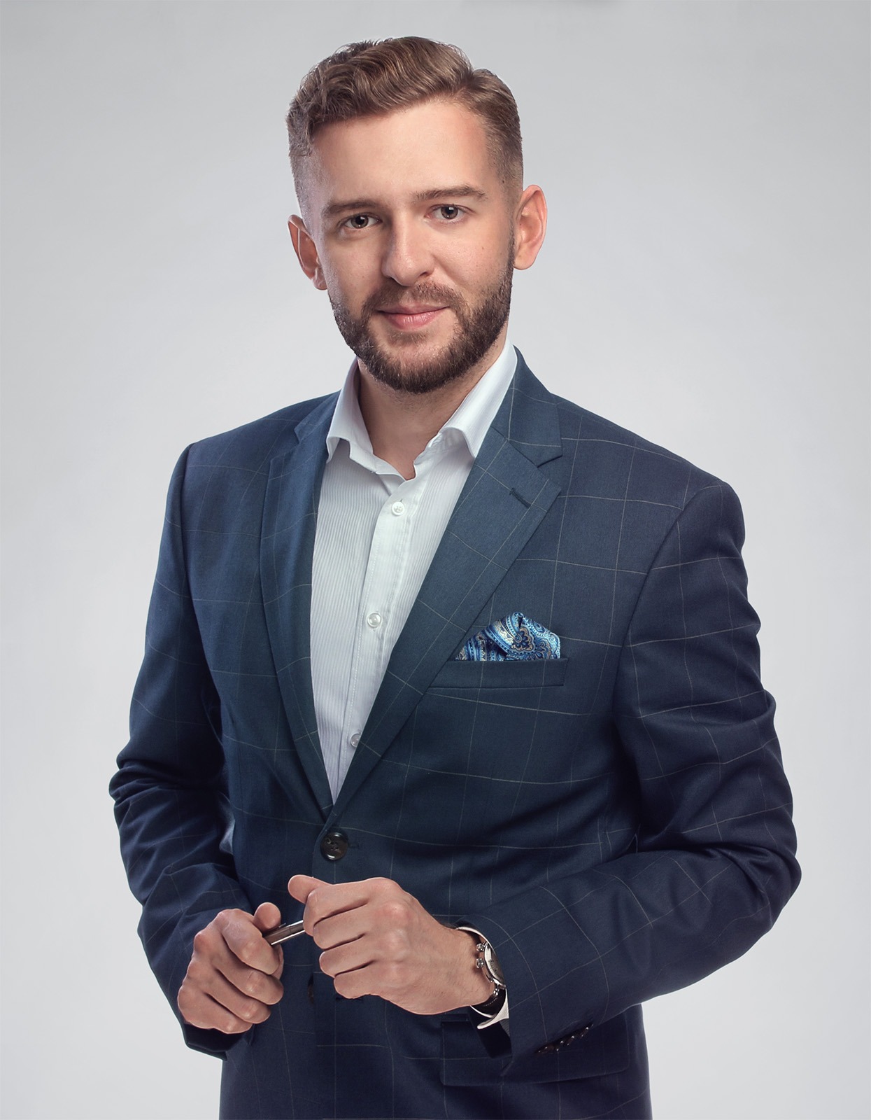 Portret uśmiechniętego mężczyzny w granatowym garniturze w kratę z poszetką i zegarkiem, trzymającego spinkę do mankietów na neutralnym tle.