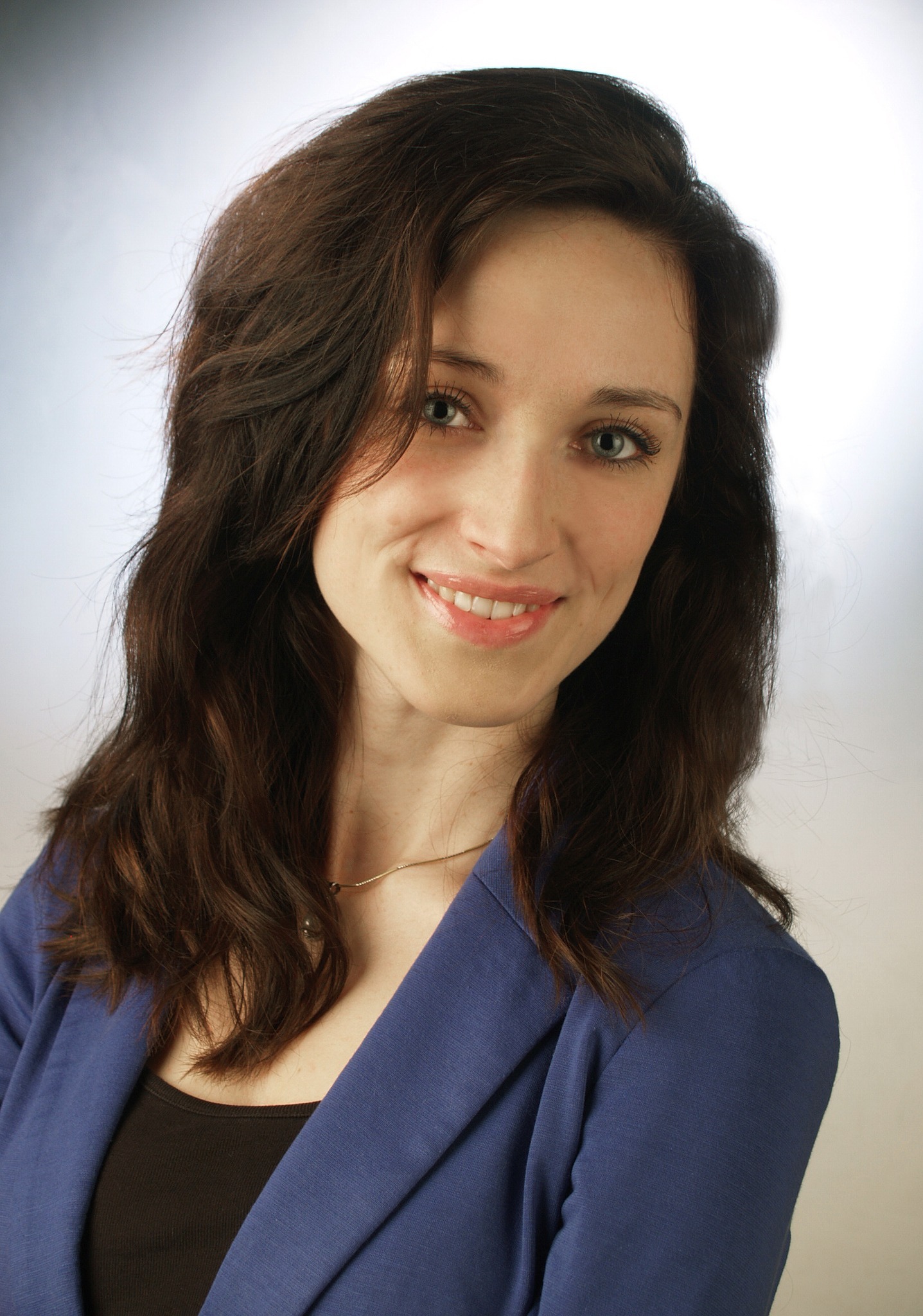 Portret uśmiechniętej kobiety w niebieskiej marynarce, z brązowymi włosami i delikatnym złotym łańcuszkiem na szyi, na rozmytym, jasnym tle.