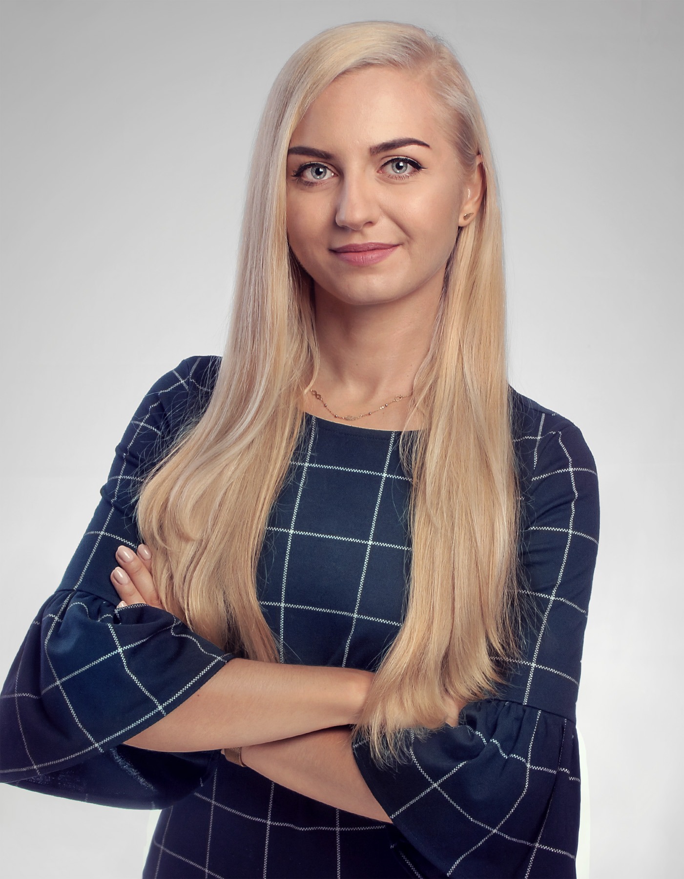Portret uśmiechniętej kobiety z blond włosami, ubranej w granatową sukienkę w kratę, z rękami skrzyżowanymi na piersiach, na jednolitym szarym tle.