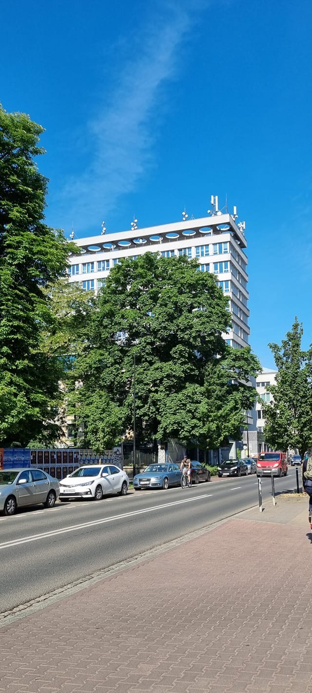 Widok na budynek biurowy z antenami na dachu, częściowo zasłonięty drzewami, z ulicą i zaparkowanymi samochodami w pierwszym planie, w słoneczny dzień.