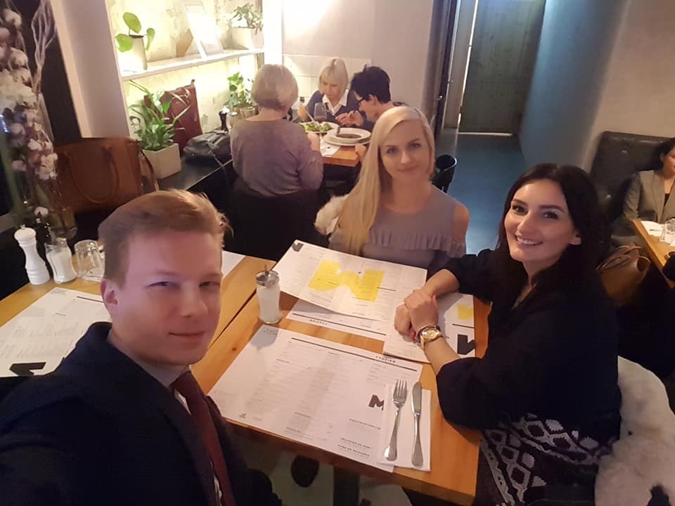 Grupa osób, w tym mężczyzna w garniturze, siedzi przy stole w restauracji, przeglądając menu i uśmiechając się do kamery.