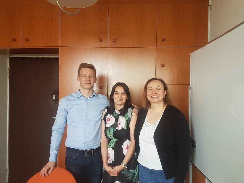 Trzy osoby (mężczyzna w niebieskiej koszuli i dwie kobiety) stoją uśmiechnięte na tle biura z drewnianymi szafkami i białą tablicą.