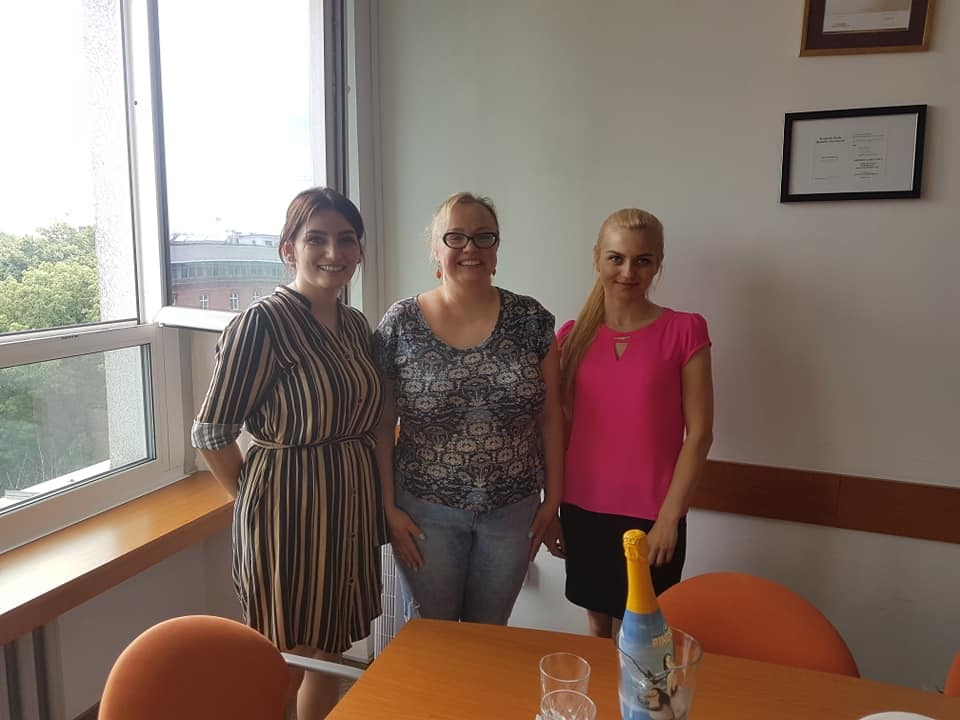 Trzy uśmiechnięte kobiety stojące w biurze, z butelką szampana na stole, w tle okno i dyplomy na ścianie.