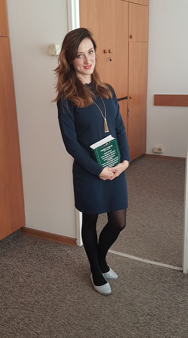 Uśmiechnięta kobieta w granatowej sukience i czarnych rajstopach, trzymająca zielony podręcznik do prawa cywilnego na tle biurowego wnętrza z drewnianymi szafkami.