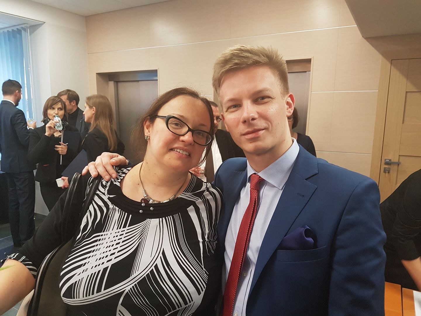 Portret kobiety w wzorzystej bluzce i mężczyzny w niebieskim garniturze z czerwoną kravatą, stojących w pomieszczeniu z innymi osobami w tle, możliwe po spotkaniu biznesowym lub konferencji.