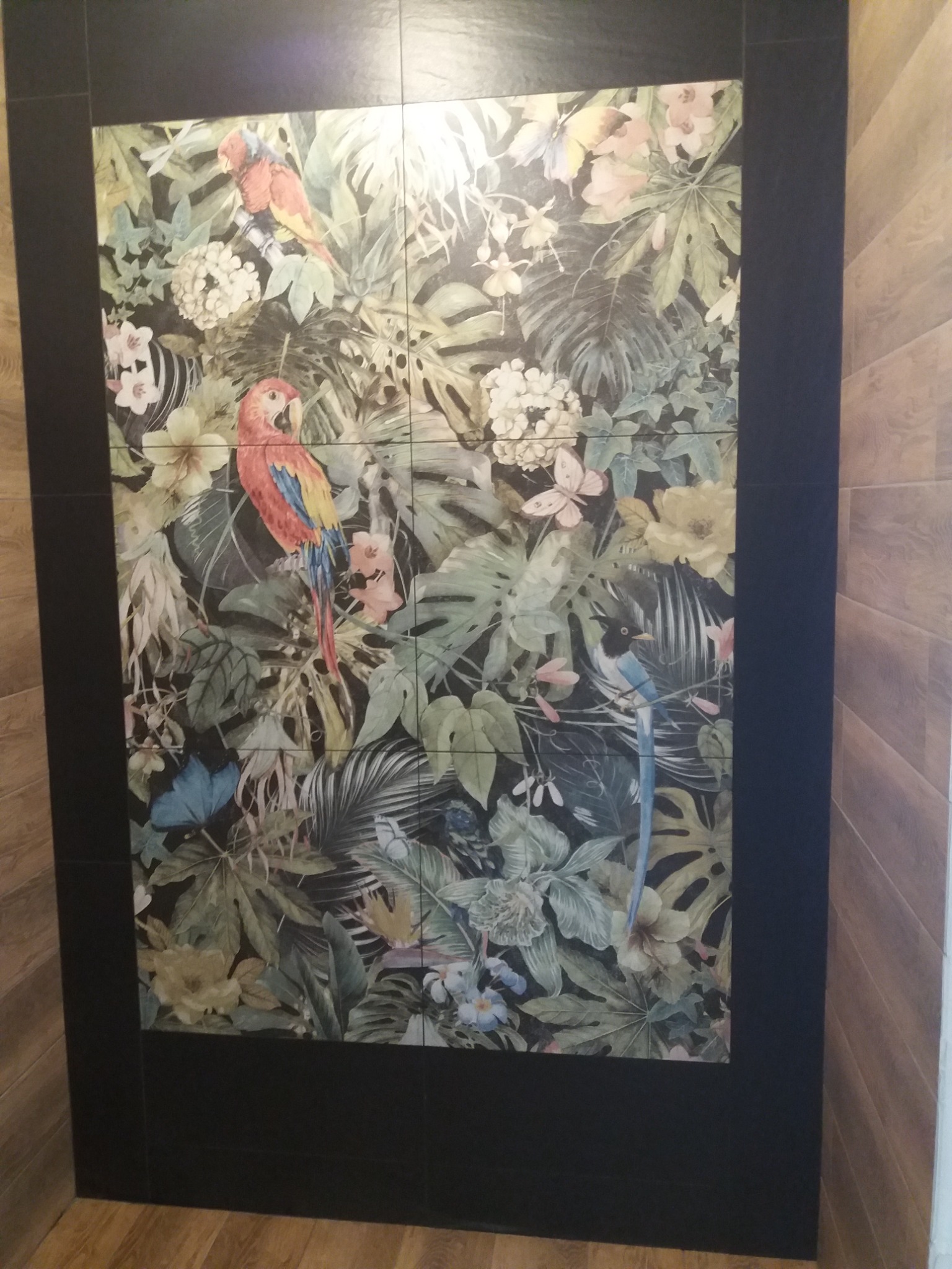 Ściana wyłożona płytkami z motywem tropikalnym: papugi, motyle i roślinność na ciemnym tle, obramowana czarnymi płytkami. Widoczny fragment drewnianej podłogi i ściany.
