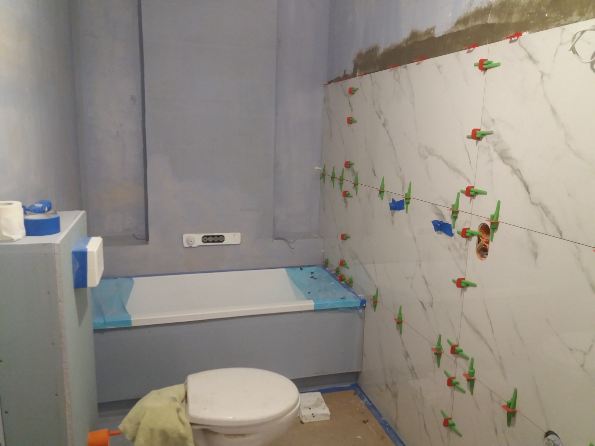 Łazienka w trakcie remontu: białe płytki imitujące marmur z systemem poziomowania, szare ściany przygotowane do malowania, zabezpieczona folią wanna oraz toaleta z ręcznikiem.