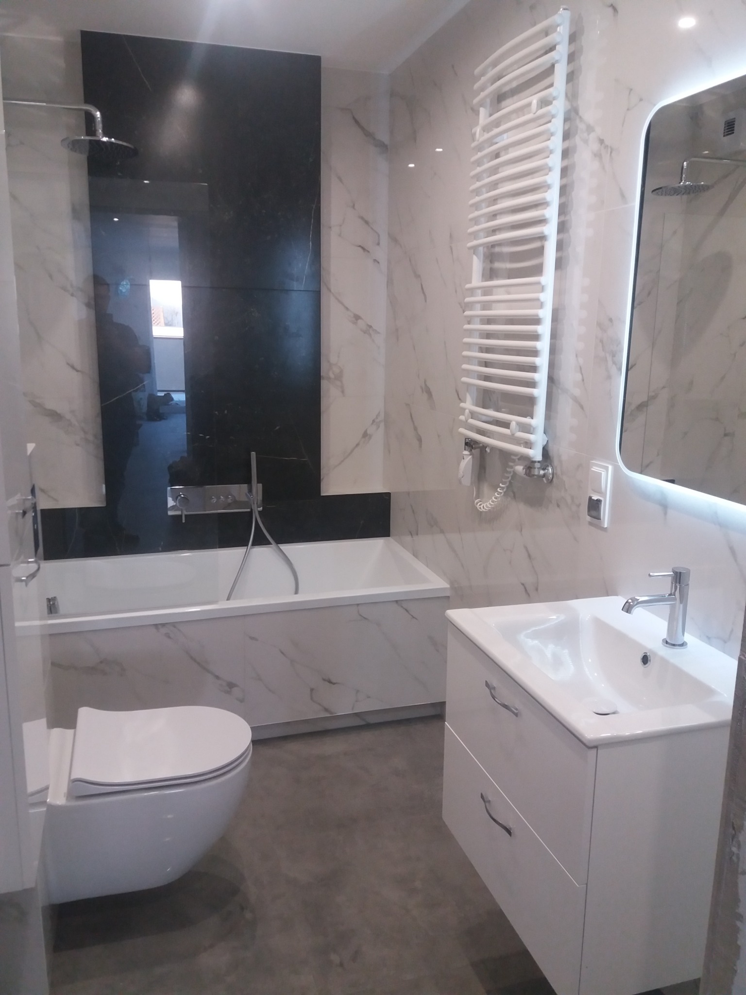 Wykończona łazienka z czarnym panelem nad wanną, białą armaturą, grzejnikiem drabinkowym i podświetlanym lustrem.