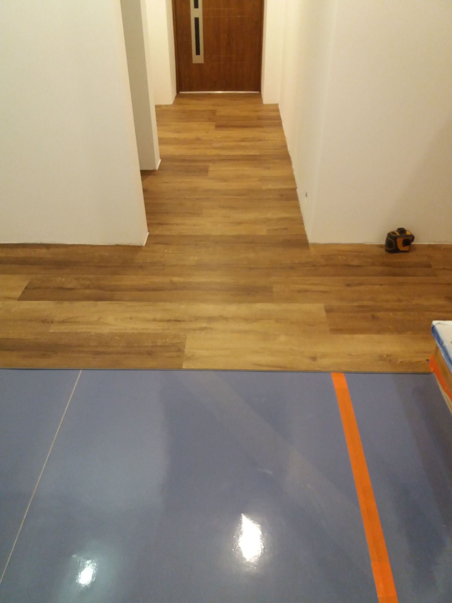 Korytarz z nową podłogą z paneli imitujących drewno, przechodzącą w niebieską posadzkę z pomarańczowym pasem, widoczny dalmierz laserowy przy ścianie.