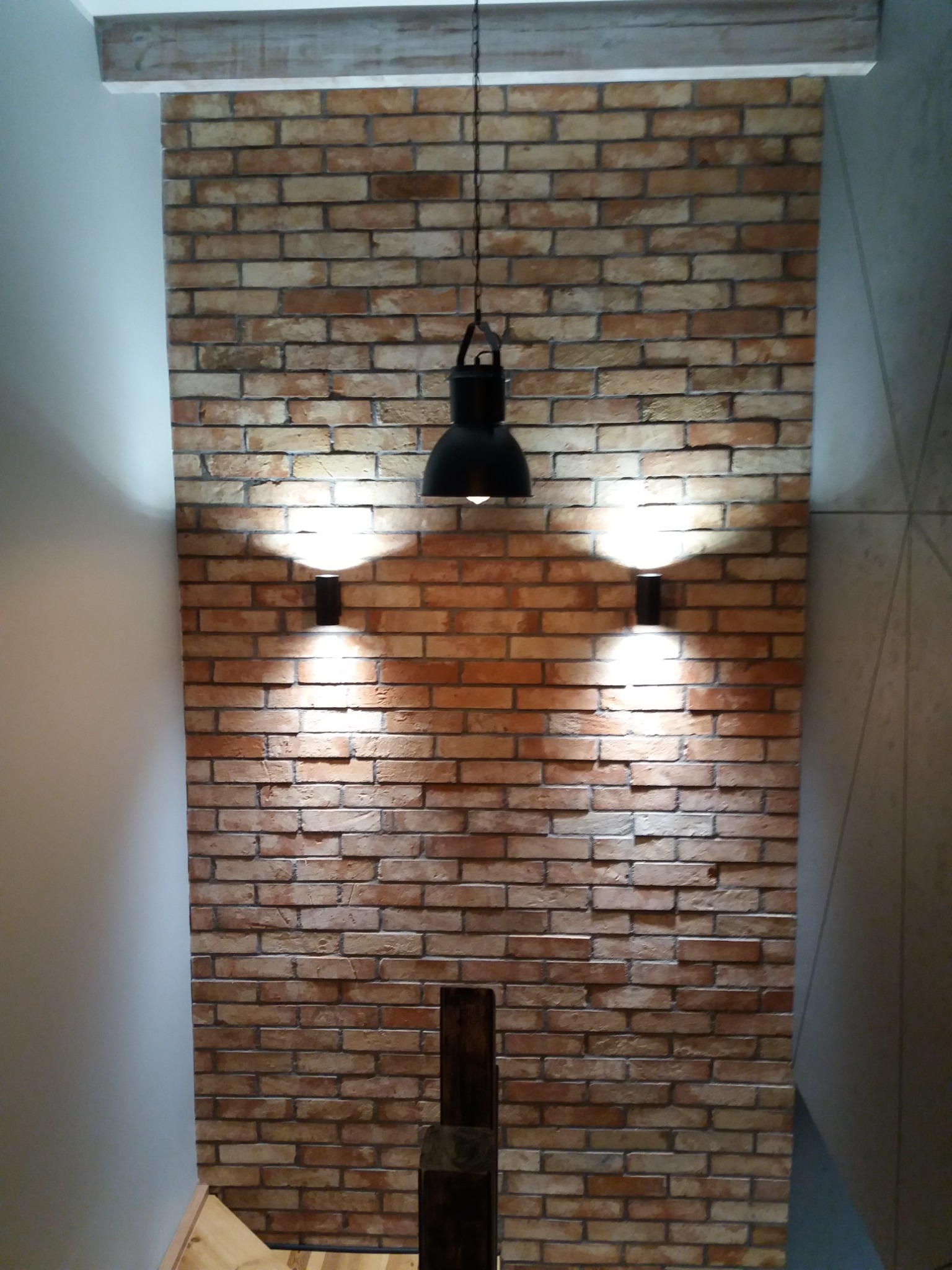 Ściana wyłożona cegłą klinkierową z zamontowanymi kinkietami i lampą wiszącą, drewniany element dekoracyjny przy suficie.