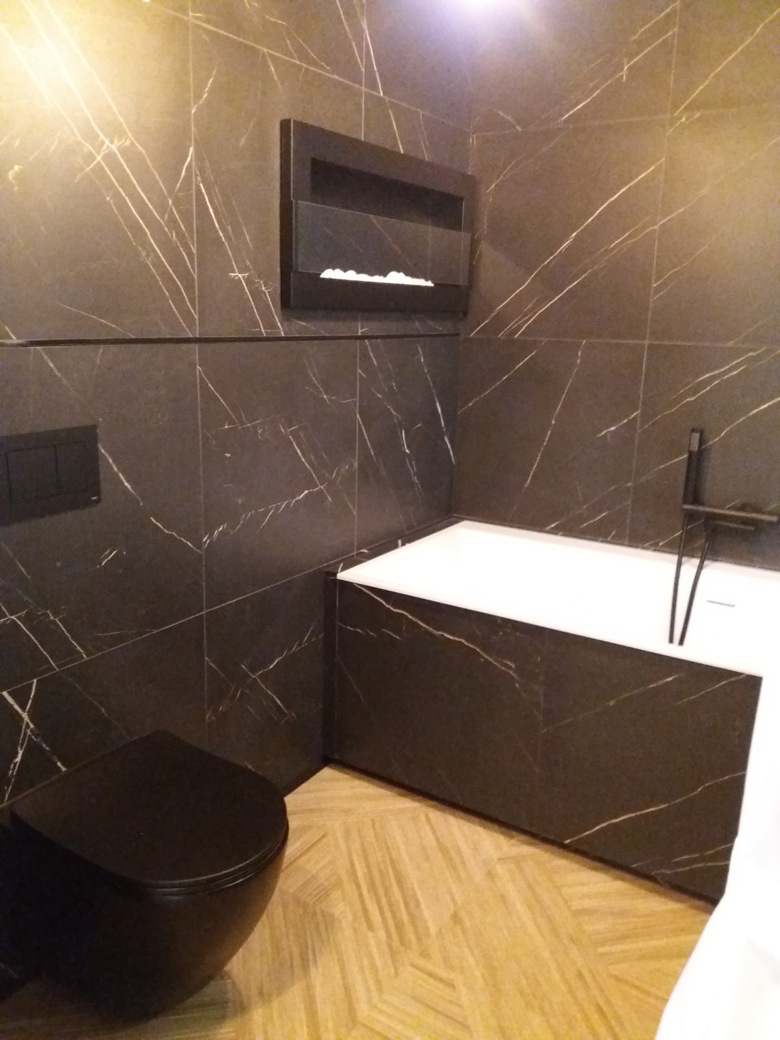 Nowoczesna łazienka z czarnymi płytkami imitującymi marmur ze złotymi żyłkami, czarną armaturą i toaletą oraz jasną drewnopodobną podłogą ułożoną w jodełkę.